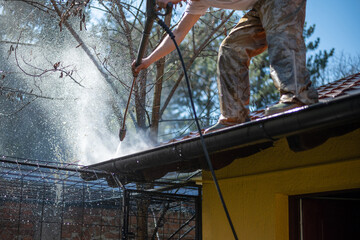 Worker standing on the roof and cleaning rain gutter with high pressure water jet. Professional...