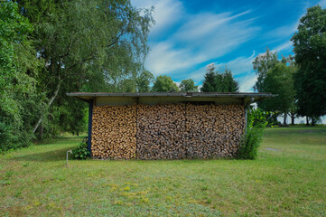 Wooden shed with stacked firewood in a meadow. Blue sky with clouds. Summertime near Berlin.