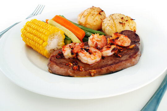 Meal setting of a steak with garlic butter sauce and fresh king prawns - surf n' turf - and fresh vegetables, on a white plate with knife and fork isolated on white