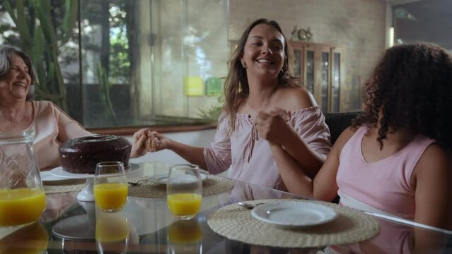 av&oacute; m&atilde;e e filha brasileiras sentadas na mesa de jantar de m&atilde;os dadas comemorando no dia das m&atilde;es