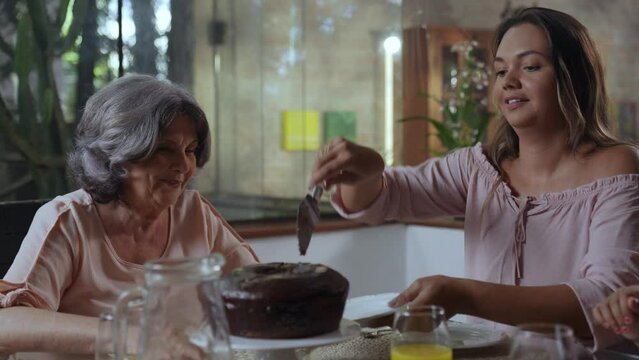 mulher brasileira sentada na mesa de jantar servindo bolo para sua m&atilde;e idosa no dia das m&atilde;es
