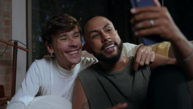 casal gay inter racial brasileiro sentado no sof&aacute; da sala fazendo uma v&iacute;deo chamada para mostrar sua alian&ccedil;a de noivado no dia dos namorados