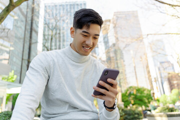 Handsome smiling asian man holding smartphone using mobile app, reading text message, communication online sitting on urban street. Happy Chinese hipster guy shopping online, ordering food on website