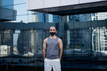 With his hands in his pockets, a toned man in athletic wear poses in an outdoor pool area. His sleeveless shirt shows his physique, and his handsome Middle Eastern features add interest.