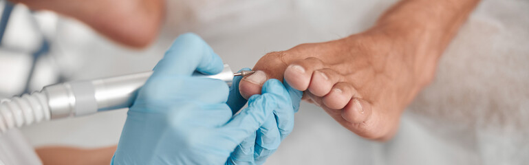Perdicure master polishes toenail of client with grinding tool at procedure in salon