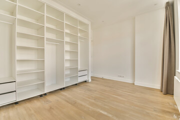 an empty room with white shelvings and wooden flooring in the middle part of the room, there is a large window