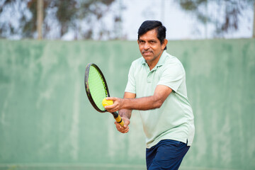 Indian senior man servicing by looking opponent at tennis court - concept of active healthy lifestyle, fitness and training.