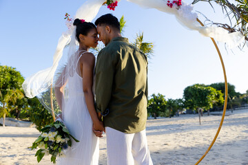 Caucasian newlywed couple with face to face holding hands and standing at beach wedding ceremony