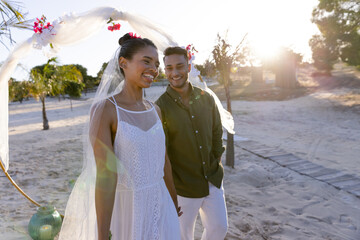Smiling caucasian newlywed couple standing at beach at wedding ceremony against sky at sunset