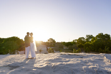 Caucasian romantic newlywed couple standing on sandy beach against clear sky at sunset, copy space