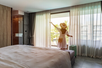 Young woman traveler opening the curtains and looking at the view from the window of a hotel room while on summer vacation, Travel lifestyle concept