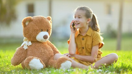 Pretty child girl passionately taking on mobile phone with her friend sitting beside a teddy bear outdoors on green grass lawn. Concept of friendship values