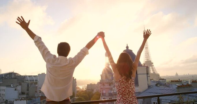 Couple, celebration and hotel balcony in Paris, together or holding hands in air, love or romantic vacation. Young man, woman and celebrate on travel, holiday or summer in France, sunset and terrace
