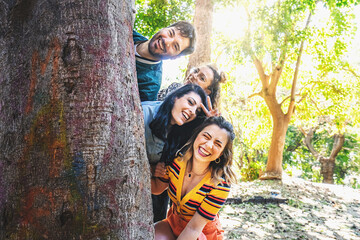 A group of friends enjoy a fun photoshoot in the park. They playfully hide their heads behind a large tree, sharing laughter and posing towards the camera. Their outfits exude a nostalgic 90s style.