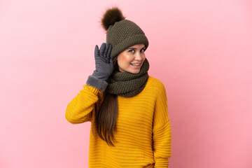 Young caucasian girl with winter hat isolated on pink background listening to something by putting hand on the ear