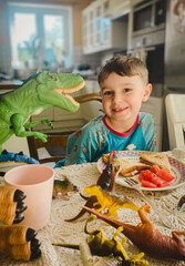 Uśmiechnięty chłopiec przy stole w kuchni, śniadanie w domu. Dinozaury na stole, zabawa przy śniadaniu. © malgo_walko
