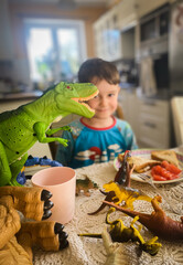 Uśmiechnięty chłopiec przy stole w kuchni, śniadanie w domu. Dinozaury na stole, zabawa przy...