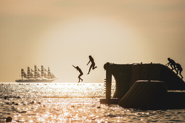 Silouettes of kids jumping in the water, with ship in the background.