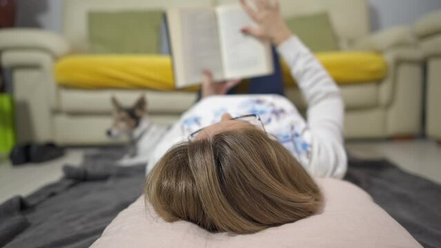 Woman lying on the floor on a blanket with her dog while turning the pages of a book.