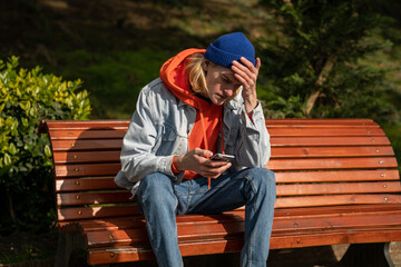 Bad news. Upset millennial guy sitting on bench in park holding smartphone reading unpleasant...
