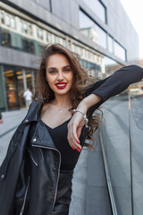 Fototapeta na wymiar Happy beautiful young fashionable girl with red lips and a smile in black elegant clothes with a leather black jacket walks and poses in the city