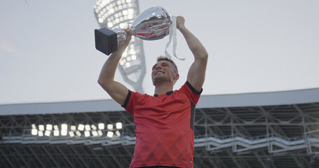 Portrait of Caucasian male soccer football player celebrating victory in the championship, lifting the trophy above his head in a huge stadium
