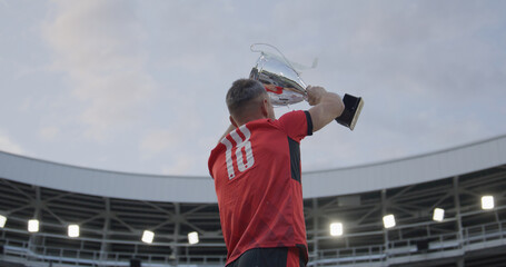 Portrait of Caucasian male soccer football player celebrating victory in the championship, lifting the trophy above his head in a huge stadium