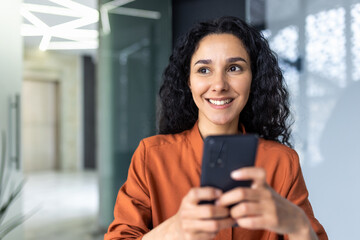 Fototapeta na wymiar Beautiful and successful Hispanic woman at work smiling and looking at the phone screen, businesswoman in the office holds smartphone in her hands closeup browses the Internet uses application call.