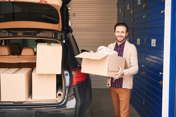 Happy man with cardboard box near the trunk of the car in a warehouse