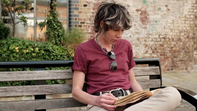Non-binary person reading a book on a bench.