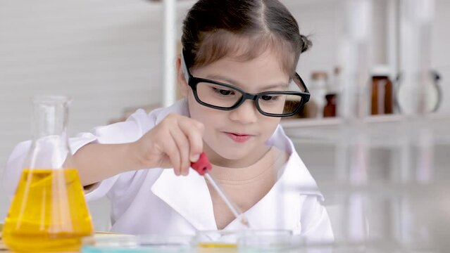 Cute little girl wearing lab coat learning chemistry in school laboratory, doing a chemical experiment while making analyzing and mixing liquid in glass at science class on the table.