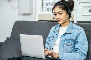 Asian young business woman is sitting on the sofa in her own house is talking in a video conference on line with a headset with microphone and laptop in home, work from home