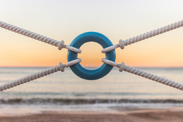 Fence of the Pedestrian way of Salinas, Asturias, with white sailboat ropes and a blue life ring, and at the background a blurry sand beach with quiet waves from the Atlantic Ocean at sundown