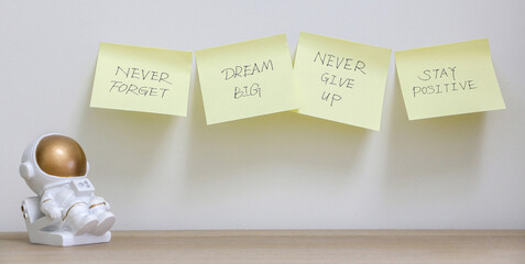 Inspiration concept, four yellow sticky notes stuck across wall with motivational messages to never forget to dream big, never give up and stay positive, besides astronaut figurine