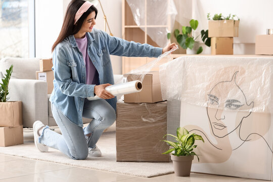 Young woman wrapping cardboard box with stretch film at home on moving day