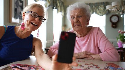 Happy smiling, laughing elderly senior woman sitting at table with mature middle aged daughter, video chat on phone. Smiling blonde lady listening to family on smartphone.
