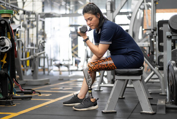 Fototapeta na wymiar Person with a limb disability doing physical exercise by weightlifting in gym. Asian bionic woman with prosthetic leg sitting workout using dumbbell in fitness studio. Healthy lifestyle female amputee