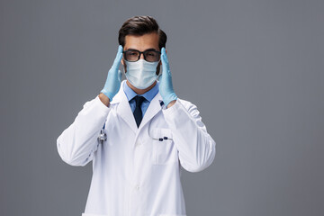 Male doctor in a white coat and medical mask and sterile gloves, eyeglasses looks at the camera on a gray isolated background, copy space, space for text, health