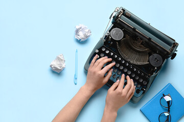 Woman typing on typewriter with crumpled paper, notebook and eyeglasses on light blue background