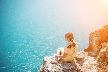 Happy girl perched atop a high rock above the sea, wearing a yellow jumpsuit and braided hair,...