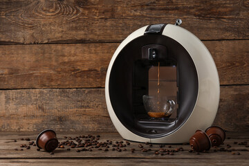 Modern capsule coffee maker with dripping espresso, cup, pods and beans on wooden table