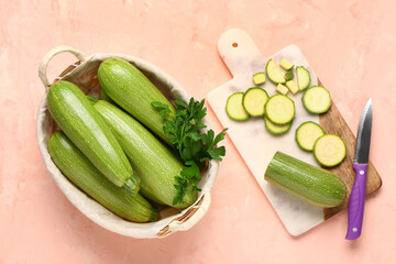 Wicker basket with fresh green zucchini and board of slices on pink background