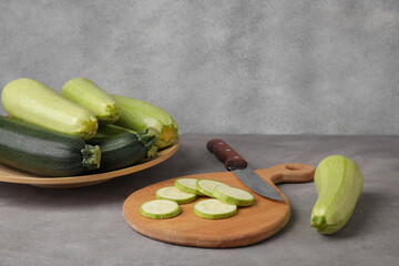 Board with slices and plate of fresh green zucchini on grey background