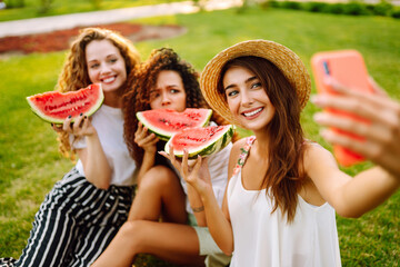 Three young woman relaxing on the grass, eating watermelon and taking selfie on the phone. Summer concept.