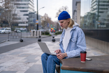 Focused young man 20s wearing denim clothes and beanie hat sit on bench with coffee cup working online on laptop. Guy student sits on city street studying online. Urban lifestyle and freelancing