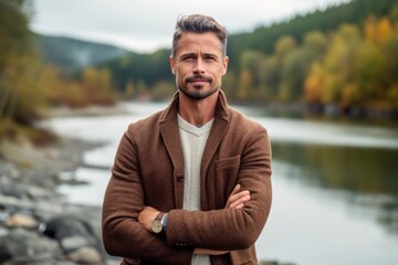 Fototapeta premium Handsome man in a brown coat with crossed arms on the background of a mountain river