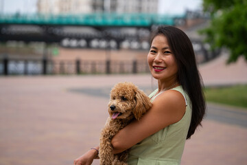 A beautiful Asian girl (Kazakh) holds a dog (mini poodle) in her hands. Summer portrait of a young woman in the park.