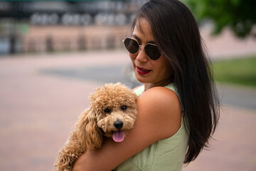 A beautiful Asian girl (Kazakh) holds a dog (mini poodle) in her hands. Summer portrait of a young woman in the park.