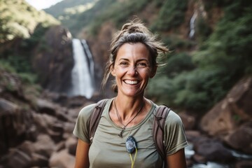 Portrait of smiling woman with backpack standing in front of waterfall in mountains