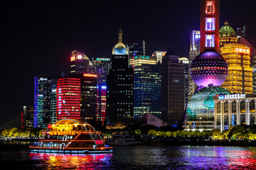 Tour boat cruises along the Huangpu River passing many spectacular modern buildings of Shanghai Lujiazui lit up at night with vibrant colours